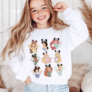 May include: White sweatshirt featuring cartoon illustrations of various princesses wearing Minnie Mouse ears. The princesses are depicted in different poses and outfits. The sweatshirt is a casual, comfortable style.