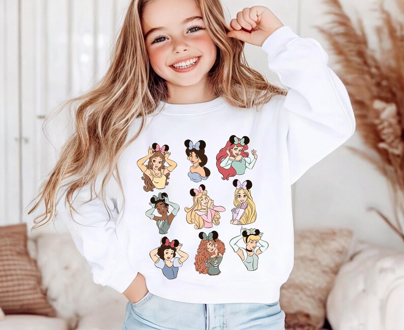 May include: White sweatshirt featuring cartoon illustrations of various princesses wearing Minnie Mouse ears. The princesses are depicted in different poses and outfits. The sweatshirt is a casual, comfortable style.