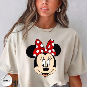Op de afbeelding: Ivoor kleurig T-shirt met een Minnie Mouse afbeelding. De afbeelding toont Minnie's gezicht met zwarte oren, een rode strik met witte stippen en een lachende uitdrukking. Het T-shirt is een casual stijl.