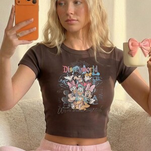 May include: A brown crop top featuring a Disney World graphic with various cartoon characters. The shirt has short sleeves and a crew neck. The text "Where dreams come true" is also visible.