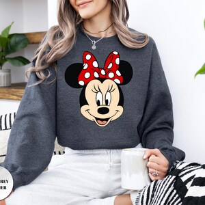 May include: Dark grey heather sweatshirt with a Minnie Mouse graphic. The graphic shows Minnie's face with a red bow with white polka dots. The sweatshirt is a crew neck style and has long sleeves. The text "DARK GREY HEATHER" is in the bottom left corner.
