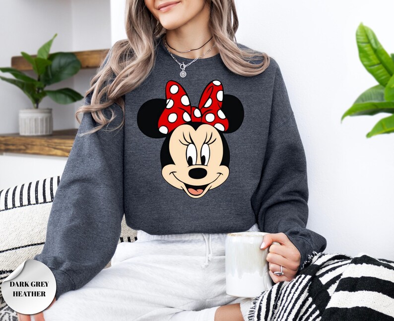 May include: Dark grey heather sweatshirt with a Minnie Mouse graphic. The graphic shows Minnie's face with a red bow with white polka dots. The sweatshirt is a crew neck style and has long sleeves. The text "DARK GREY HEATHER" is in the bottom left corner.