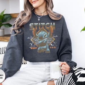 May include: A dark grey heather long-sleeve sweatshirt featuring a graphic of the blue character Stitch with arms crossed. The design includes the text STITCH and EXP 626 in a distressed orange font, set against a geometric diamond background.