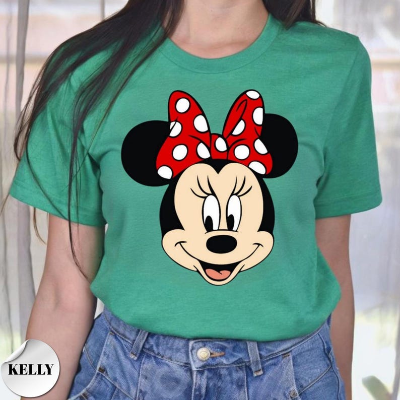 Puede incluir: Camiseta verde con un gr&aacute;fico de Minnie Mouse. El personaje de dibujos animados tiene una cara negra, orejas grandes y negras y un lazo rojo con lunares blancos. La camiseta es de color verde liso.