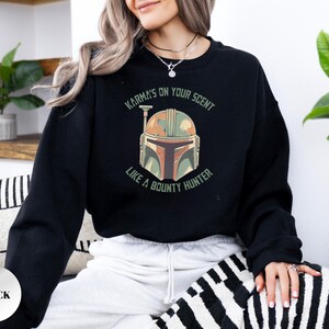 Puede incluir: Una sudadera negra de manga larga con un estampado gr&aacute;fico de un casco en tonos verdes, naranjas y marrones. El texto que rodea el casco dice, Karma's on your scent like a bounty hunter, impreso en una fuente verde oscuro sin remates.