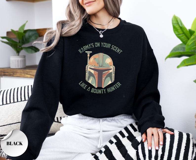 Puede incluir: Una sudadera negra de manga larga con un estampado gr&aacute;fico de un casco en tonos verdes, naranjas y marrones. El texto que rodea el casco dice, Karma's on your scent like a bounty hunter, impreso en una fuente verde oscuro sin remates.