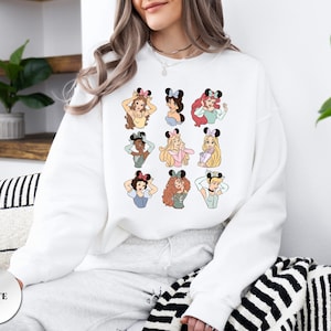 May include: White sweatshirt featuring a colorful graphic of cartoon princesses wearing mouse ears. The princesses are in various poses and have different hairstyles and skin tones. The word "WHITE" is printed on a white circle on the bottom left.