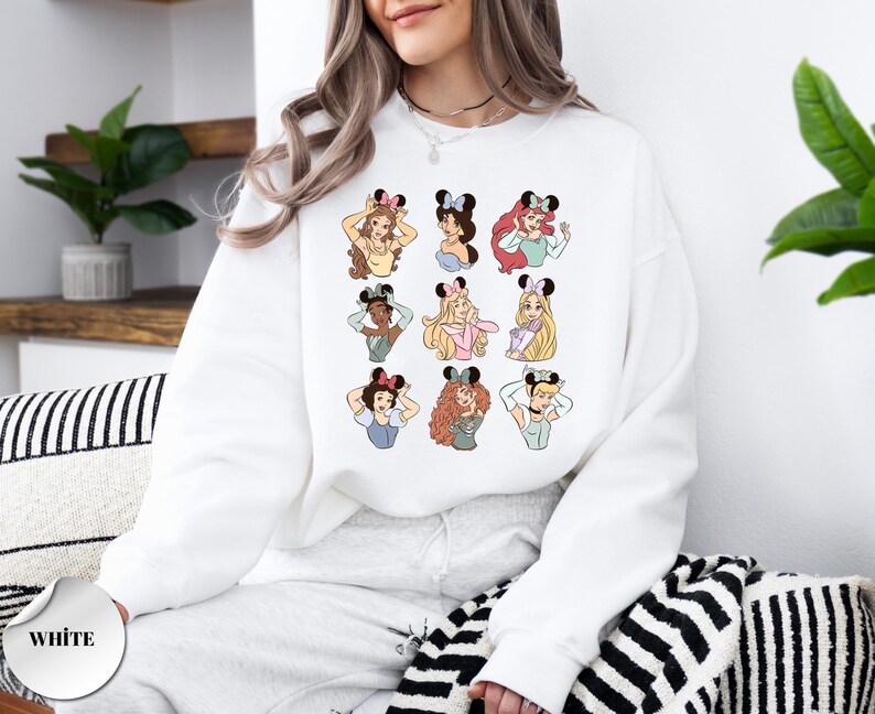 May include: White sweatshirt featuring a colorful graphic of cartoon princesses wearing mouse ears. The princesses are in various poses and have different hairstyles and skin tones. The word "WHITE" is printed on a white circle on the bottom left.