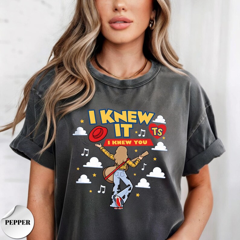 Puede incluir: Una camiseta gris carb&oacute;n de manga corta con un estampado gr&aacute;fico de una persona de pelo largo tocando la guitarra. El texto dice I KNEW IT e I KNEW YOU, acompa&ntilde;ado de un sombrero rojo, un coraz&oacute;n rojo con TS, notas musicales, estrellas y nubes.