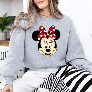 Puede incluir: Una sudadera gris claro con un gr&aacute;fico de Minnie Mouse. El gr&aacute;fico muestra la cara de Minnie con un lazo rojo con lunares blancos. La palabra "GREY" est&aacute; impresa en un c&iacute;rculo gris en la esquina inferior izquierda. La sudadera es de estilo casual.
