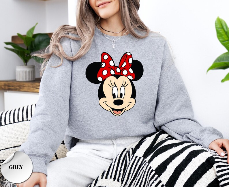 Puede incluir: Una sudadera gris claro con un gr&aacute;fico de Minnie Mouse. El gr&aacute;fico muestra la cara de Minnie con un lazo rojo con lunares blancos. La palabra "GREY" est&aacute; impresa en un c&iacute;rculo gris en la esquina inferior izquierda. La sudadera es de estilo casual.