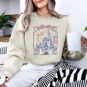 May include: Sand-colored sweatshirt featuring a whimsical castle illustration with a fairy, fireworks, and pastel balloons. The design is framed by a pink ribbon and floral accents. The sweatshirt is a relaxed fit.
