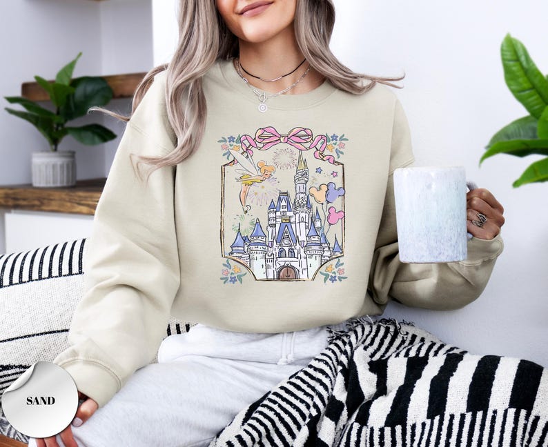 May include: Sand-colored sweatshirt featuring a whimsical castle illustration with a fairy, fireworks, and pastel balloons. The design is framed by a pink ribbon and floral accents. The sweatshirt is a relaxed fit.