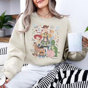 May include: A sand-colored crewneck sweatshirt featuring a graphic print of Toy Story characters including Woody, Buzz Lightyear, Jessie, Rex, Hamm, Mr. Potato Head, Bullseye, Bo Peep, and Forky, with their names written in a cursive script font.
