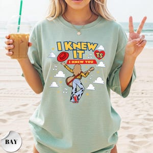 Puede incluir: Una camiseta verde salvia con un estampado gr&aacute;fico de una persona de pelo largo tocando la guitarra, con camisa amarilla y pantalones con estampado de vaca. El texto dice I KNEW IT e I KNEW YOU, junto a un sombrero rojo, un coraz&oacute;n rojo, nubes y notas musicales.