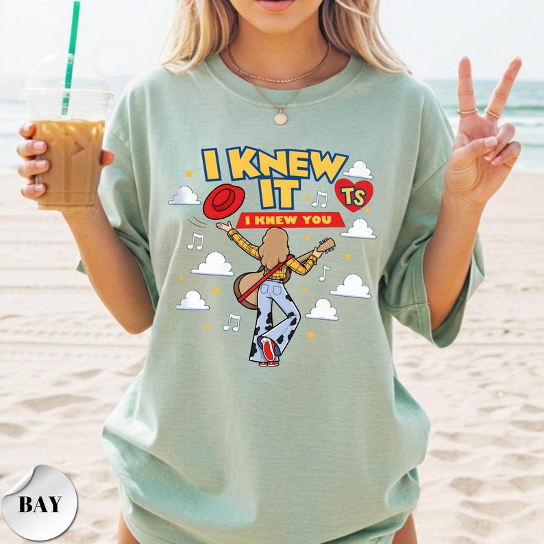 Puede incluir: Una camiseta verde salvia con un estampado gr&aacute;fico de una persona de pelo largo tocando la guitarra, con camisa amarilla y pantalones con estampado de vaca. El texto dice I KNEW IT e I KNEW YOU, junto a un sombrero rojo, un coraz&oacute;n rojo, nubes y notas musicales.