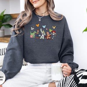 Op de afbeelding: Een donkergrijs gem&ecirc;leerd sweatshirt met ronde hals en een grafische print van diverse geanimeerde speelgoedfiguren, waaronder een cowboy, een space ranger, een cowgirl, een dinosaurus en een varken, met kleine muisoorvormige iconen.