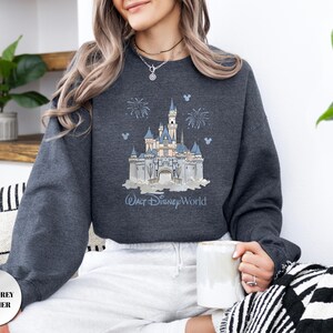 May include: A dark grey heather sweatshirt featuring a graphic print of a light blue and white castle with fireworks and small Mickey Mouse head silhouettes. The text Walt Disney World is printed in blue script below the castle illustration.