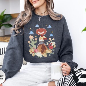 K&ouml;nnte beinhalten: Ein dunkelgrau meliertes Sweatshirt mit Rundhalsausschnitt und einem Grafikdruck einer Cowgirl mit rotem Hut auf einem braunen Pferd, umgeben von bunten W&uuml;stenblumen, Kakteen, kleinen blauen Wolken und gelben Sternen auf dunkelgrauem Grund.
