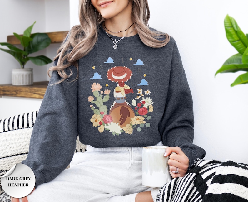 K&ouml;nnte beinhalten: Ein dunkelgrau meliertes Sweatshirt mit Rundhalsausschnitt und einem Grafikdruck einer Cowgirl mit rotem Hut auf einem braunen Pferd, umgeben von bunten W&uuml;stenblumen, Kakteen, kleinen blauen Wolken und gelben Sternen auf dunkelgrauem Grund.
