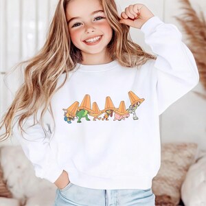 Op de afbeelding: Witte sweatshirt met een cartoonontwerp van personages die oranje verkeerskegelhoeden dragen. De afbeelding bevat een groene dinosaurus en andere figuren. De sweatshirt is een casual stijl.
