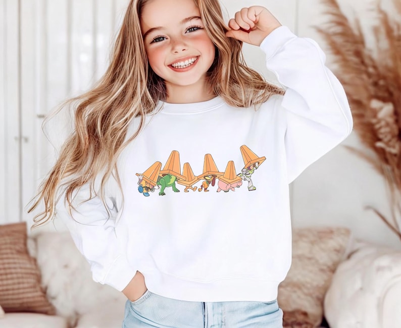Op de afbeelding: Witte sweatshirt met een cartoonontwerp van personages die oranje verkeerskegelhoeden dragen. De afbeelding bevat een groene dinosaurus en andere figuren. De sweatshirt is een casual stijl.