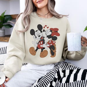 May include: Sand-colored sweatshirt featuring a graphic of Mickey and Minnie Mouse embracing, with hearts above them. Mickey is in red shorts and brown shoes, Minnie in a red polka-dot skirt and bow. The sweatshirt is a casual style.