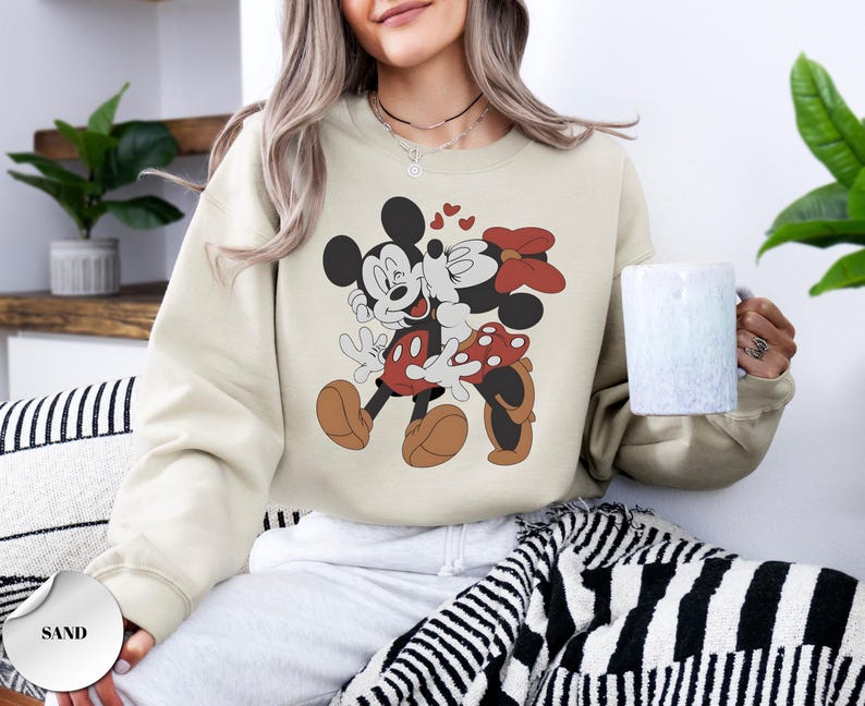 May include: Sand-colored sweatshirt featuring a graphic of Mickey and Minnie Mouse embracing, with hearts above them. Mickey is in red shorts and brown shoes, Minnie in a red polka-dot skirt and bow. The sweatshirt is a casual style.