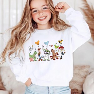 Op de afbeelding: Een wit sweatshirt met lange mouwen en een ronde hals, voorzien van een kleurrijke grafische print van diverse geanimeerde personages uit de Toy Story-franchise, waaronder Woody, Buzz Lightyear, Jessie, Rex, Bullseye en Hamm, met Mickey Mouse-ballonnen.