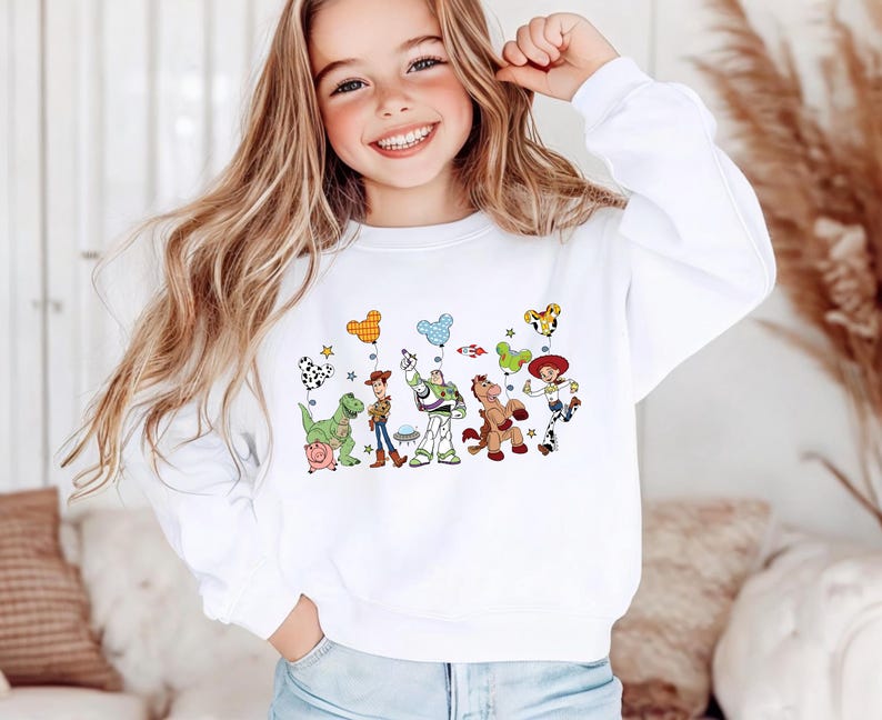 Op de afbeelding: Een wit sweatshirt met lange mouwen en een ronde hals, voorzien van een kleurrijke grafische print van diverse geanimeerde personages uit de Toy Story-franchise, waaronder Woody, Buzz Lightyear, Jessie, Rex, Bullseye en Hamm, met Mickey Mouse-ballonnen.