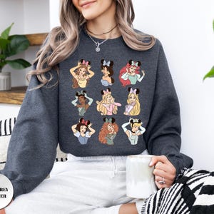 May include: Dark grey heather sweatshirt featuring a colorful graphic of Disney princesses wearing Minnie Mouse ears. The sweatshirt has the text "DARK GREY HEATHER" at the bottom left. The design includes various princesses with different hairstyles and outfits.