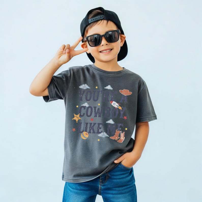 May include: A charcoal gray short-sleeve t-shirt featuring the text "YOU'RE A COWBOY LIKE ME" in dark blue letters, surrounded by graphic illustrations of a rocket, a cowboy hat, cowboy boots, stars, clouds, and a ball on a light background.