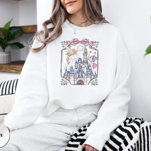 May include: White sweatshirt featuring a colorful illustration of a fairytale castle with fireworks, balloons, and a fairy. The design is framed by a pink bow and floral accents. The word "WHITE" is printed on a white circle.
