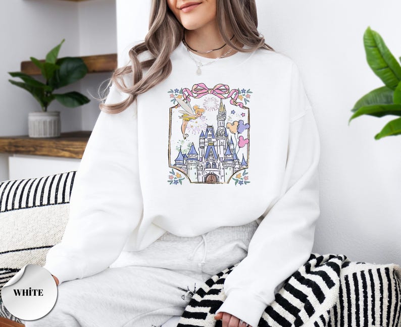 May include: White sweatshirt featuring a colorful illustration of a fairytale castle with fireworks, balloons, and a fairy. The design is framed by a pink bow and floral accents. The word "WHITE" is printed on a white circle.