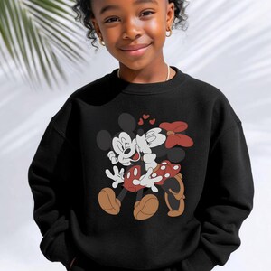 May include: Black sweatshirt featuring a graphic of Mickey Mouse and Minnie Mouse embracing, with hearts above them. Mickey is in white gloves, red shorts, and brown shoes. Minnie wears a red bow and a polka-dot skirt. The sweatshirt is worn by a young person.
