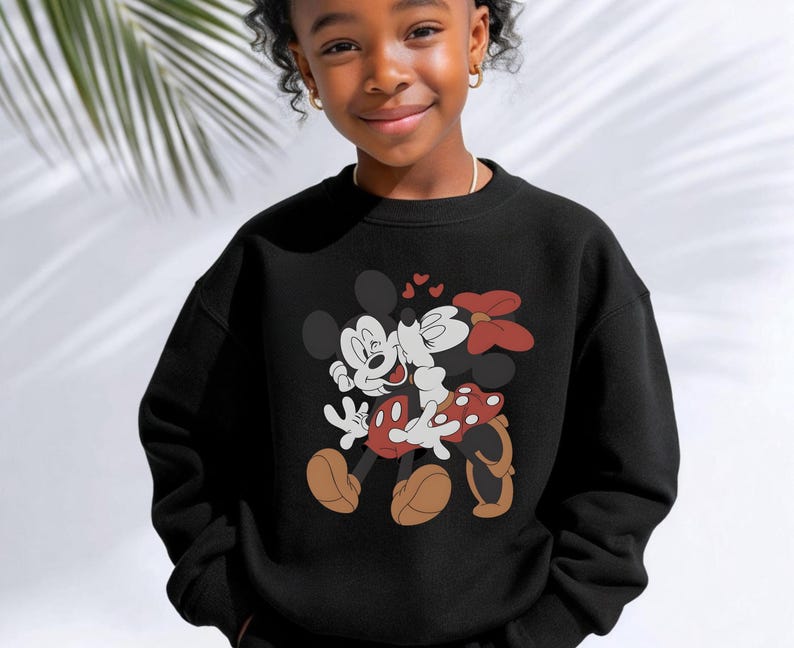 May include: Black sweatshirt featuring a graphic of Mickey Mouse and Minnie Mouse embracing, with hearts above them. Mickey is in white gloves, red shorts, and brown shoes. Minnie wears a red bow and a polka-dot skirt. The sweatshirt is worn by a young person.