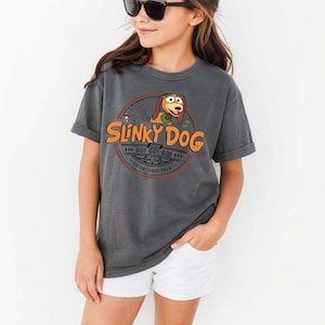 以下が含まれることがあります： チャコールグレーの半袖Tシャツ。スリンキードッグの漫画風イラストが描かれた円形のグラフィックが特徴です。テキストには「Slinky Dog Dash」、「Go! Go! Go!」、「Established 2018」と記されており、オレンジ色の文字と茶色の犬のイラストがデザインされています。