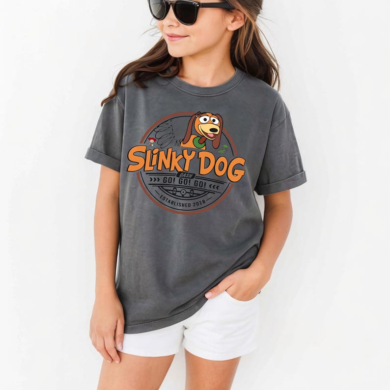 以下が含まれることがあります： チャコールグレーの半袖Tシャツ。スリンキードッグの漫画風イラストが描かれた円形のグラフィックが特徴です。テキストには「Slinky Dog Dash」、「Go! Go! Go!」、「Established 2018」と記されており、オレンジ色の文字と茶色の犬のイラストがデザインされています。