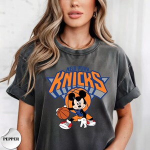 Puede incluir: Una camiseta de manga corta color gris carb&oacute;n con un estampado gr&aacute;fico de Mickey Mouse vistiendo un uniforme de baloncesto y sosteniendo un bal&oacute;n. Sobre el personaje, el texto NEW YORK KNICKS aparece en letras naranjas con bordes azules.