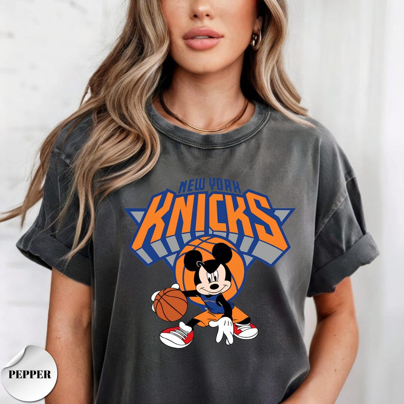 Puede incluir: Una camiseta de manga corta color gris carb&oacute;n con un estampado gr&aacute;fico de Mickey Mouse vistiendo un uniforme de baloncesto y sosteniendo un bal&oacute;n. Sobre el personaje, el texto NEW YORK KNICKS aparece en letras naranjas con bordes azules.