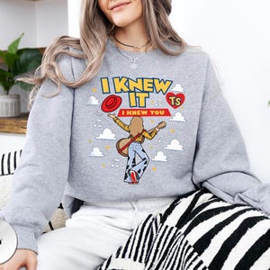 May include: A grey crewneck sweatshirt featuring a graphic print of a person playing an acoustic guitar. The text reads I KNEW IT and I KNEW YOU, accompanied by a red hat, a red heart with TS, musical notes, and white clouds on a grey background.