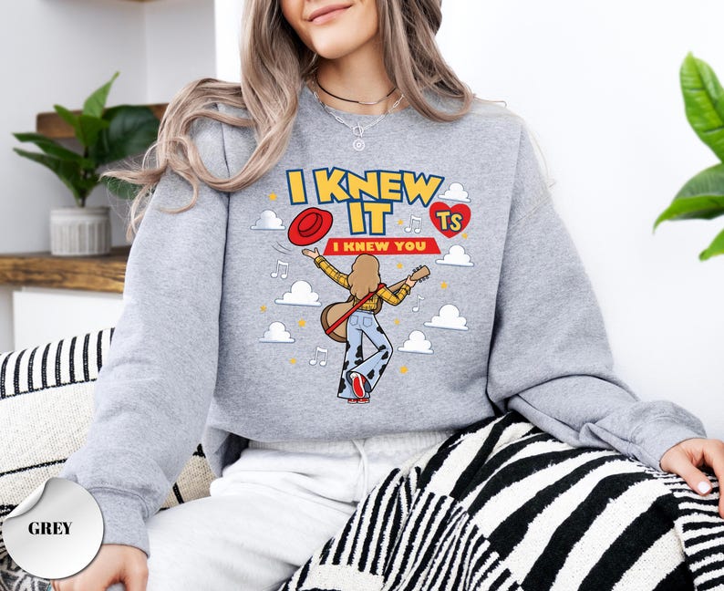 May include: A grey crewneck sweatshirt featuring a graphic print of a person playing an acoustic guitar. The text reads I KNEW IT and I KNEW YOU, accompanied by a red hat, a red heart with TS, musical notes, and white clouds on a grey background.