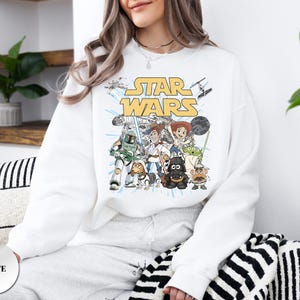 Puede incluir: Una sudadera blanca de manga larga con un estampado gr&aacute;fico del texto Star Wars en amarillo sobre un grupo de personajes de dibujos animados vestidos como figuras de Star Wars, incluyendo un vaquero, una vaquera, un perro y un alien&iacute;gena.