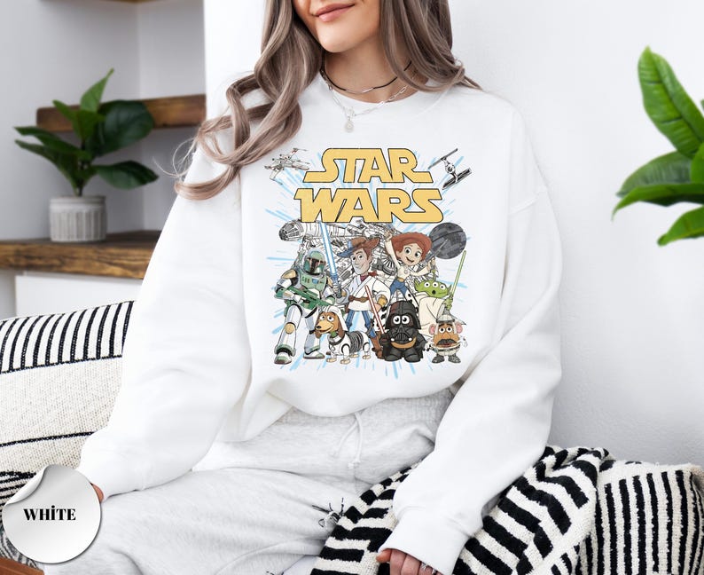 Puede incluir: Una sudadera blanca de manga larga con un estampado gr&aacute;fico del texto Star Wars en amarillo sobre un grupo de personajes de dibujos animados vestidos como figuras de Star Wars, incluyendo un vaquero, una vaquera, un perro y un alien&iacute;gena.