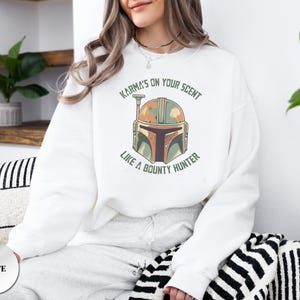 Puede incluir: Una sudadera blanca de manga larga con un gr&aacute;fico de un casco estilizado en tonos naranja, verde y marr&oacute;n. El texto "KARMA'S ON YOUR SCENT LIKE A BOUNTY HUNTER" est&aacute; impreso en letras may&uacute;sculas verdes en un arco alrededor del casco.