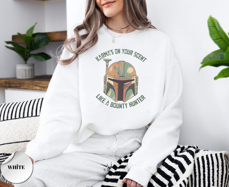Puede incluir: Una sudadera blanca de manga larga con un gr&aacute;fico de un casco estilizado en tonos naranja, verde y marr&oacute;n. El texto "KARMA'S ON YOUR SCENT LIKE A BOUNTY HUNTER" est&aacute; impreso en letras may&uacute;sculas verdes en un arco alrededor del casco.