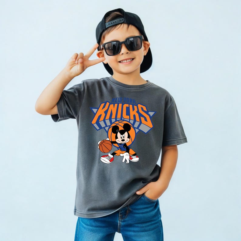 Puede incluir: Una camiseta de manga corta gris carb&oacute;n con un gr&aacute;fico de Mickey Mouse jugando al baloncesto y el texto NEW YORK KNICKS en naranja y azul. El dise&ntilde;o incluye un fondo de canasta de baloncesto y a Mickey vistiendo una camiseta azul y naranja.