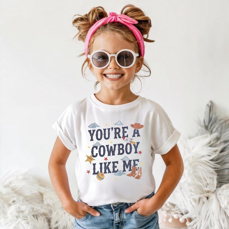 May include: A white short-sleeved t-shirt featuring the dark blue text "YOU'RE A COWBOY LIKE ME" surrounded by colorful illustrations of a rocket, a cowboy hat, a star, a ball, and boots, paired with blue denim jeans and a bright pink headband.