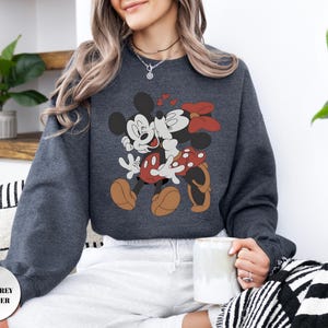 May include: Dark grey heather sweatshirt featuring a graphic of Mickey and Minnie Mouse embracing, with red hearts and bows. The cartoon characters are in their classic outfits. The sweatshirt has a crew neck and long sleeves.