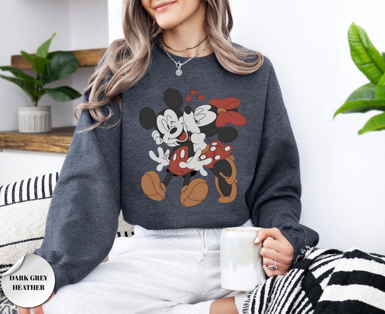 May include: Dark grey heather sweatshirt featuring a graphic of Mickey and Minnie Mouse embracing, with red hearts and bows. The cartoon characters are in their classic outfits. The sweatshirt has a crew neck and long sleeves.